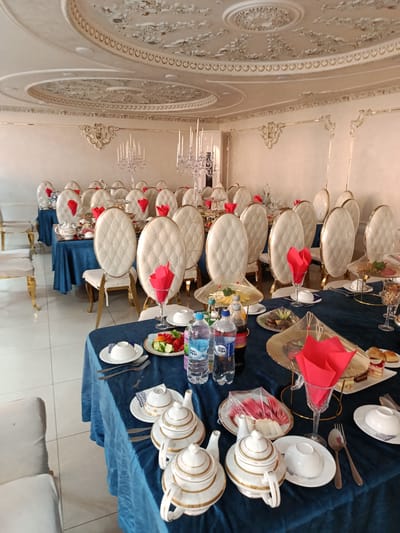The image depicts a formal dining setup, likely for an event, in a room in Shahrisabz City, Uzbekistan. Multiple tables are draped with dark blue tablecloths, each set with white crockery, silverware, and glassware. White upholstered chairs with gold accents are arranged around the tables. Red napkins are prominently displayed on each place setting. In the foreground, two teapots and cups are visible on a table laden with food, including a fruit platter and what appears to be pastries. In the background, more tables and chairs are arranged, and decorative candelabras are present. The room features ornate ceiling and wall decorations, suggesting a high-end establishment. No people are visible in the image.