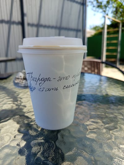 The image shows a white paper coffee cup with handwritten text on it. The cup is placed on a glass table with a textured surface, casting shadows. In the background, a blurred fence and greenery are visible. The coffee cup appears to be outdoors, possibly in a yard or patio area. The handwritten text on the cup reads: "Преграды - это повод стать сильней", which translates to "Obstacles are a reason to become stronger".