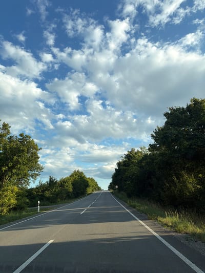 The image shows a rural asphalt road in the municipality of Veľaty, Slovakia, on a clear day with scattered white clouds against a blue sky. The road stretches into the distance, flanked by lush green trees and vegetation on both sides. White lines mark the edges and center of the road. A circular red and white traffic sign is visible on the right side of the road in the distance. The sunlight casts shadows across the asphalt, indicating the time of day is likely afternoon. There are no people or vehicles visible in the image.