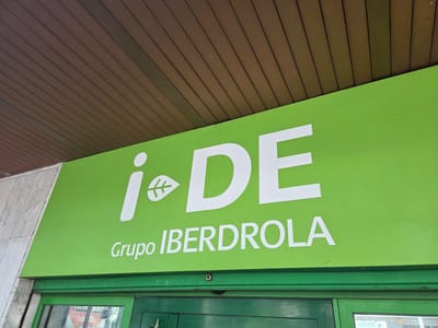 A green sign with white lettering hangs above a building entrance in Collado Villalba, Spain. The sign reads "i DE" followed by "Grupo IBERDROLA". The building has a brown slatted overhang and a facade of grey speckled stone. Below the sign, a green doorway with glass panels is visible. The image captures a daytime view of the building's exterior.
