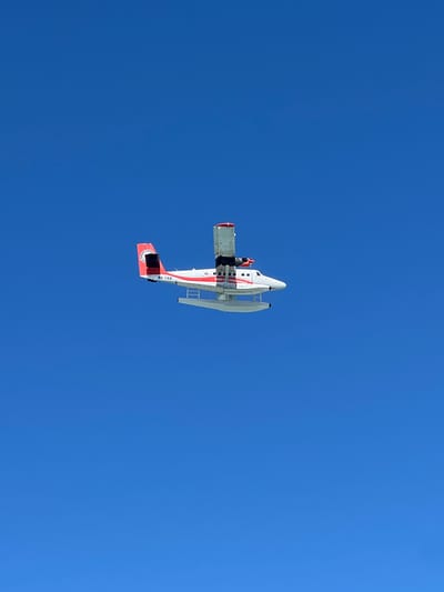 A de Havilland Canada DHC-6 Twin Otter seaplane, registered as 8Q-TAO, is captured in flight against a clear blue sky. The seaplane features a white fuselage with red accents on the tail fin and wings. The registration number "8Q-TAO" is clearly visible on the tail. The aircraft is equipped with floats for water landings and takeoffs. The image appears to have been taken from below the aircraft, looking upwards. The context provided suggests the image was taken in Malé, Maldives, a location known for its seaplane operations.