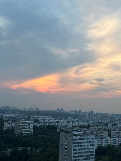The image depicts a cityscape at dusk, likely from a high vantage point. The sky is a dominant feature, displaying a gradient of colors from a soft blue at the top to warmer oranges and pinks towards the horizon, indicating the setting sun. Wispy clouds are scattered across the sky. Below the sky, a dense urban landscape unfolds, characterized by numerous apartment buildings of varying heights and architectural styles. Many buildings appear to be Soviet-era panel buildings, common in Russian cities. Lush green trees are interspersed throughout the city, particularly in the foreground and between buildings, suggesting parks or tree-lined streets. In the distance, a more distant skyline of taller buildings is visible, fading into a hazy atmosphere. There are no people or discernible actions captured in the image. The overall impression is one of a vast residential area under a dramatic sky. The description of "Borduki, Russia" is not directly ascertainable from the visual information alone, but the architectural style and urban planning are consistent with many Russian cities.