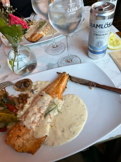 The image depicts a close-up view of a meal and drinks on a marble-patterned tabletop, likely at a restaurant in Östersund, Sweden, given the presence of a Ramlösa can. In the foreground, a white rectangular plate holds a portion of cooked salmon, generously coated in a creamy sauce, garnished with dill. To the left of the salmon, a side of sautéed vegetables including what appears to be cabbage and bell peppers, along with a single cooked mushroom, are arranged. A silver knife with a serrated edge rests on the plate to the right of the salmon.
Further back, a glass of water, or possibly a clear beverage, is positioned to the left, with faint text indicating "25 cl". To the right of this glass is a silver can of "Ramlösa ORIGINAL" mineral water, with blue and white labeling. A slice of lemon is visible next to the can. Behind the glass of water and to the left of the plate is a small, clear glass vase containing a single pink rose and some delicate white flowers, accompanied by green foliage. A fork is partially visible to the far left, resting near the vegetables. The background is out of focus, showing hints of other place settings or restaurant elements. The overall scene suggests a dining experience, with the presence of local Swedish products.