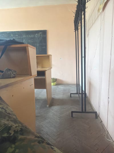 The image shows an interior space in Dolna Mitropolia, Bulgaria. In the foreground, a section of wooden shelving or cabinetry is visible, partially obscuring a blackboard mounted on the wall. The blackboard has handwritten text in a Cyrillic script, potentially containing numerals and short phrases. Above the shelving unit, a metal clothes rack runs along the length of the wall, holding several empty hangers. The floor is covered with patterned tile. A person is present in the photo with a part of the camouflage pattern.