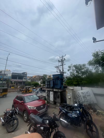 This image depicts a street scene in Kurnool, India, on a cloudy day. In the foreground, several motorcycles are parked. A dark red SUV is also present, parked on the street. Behind the vehicles, a transformer and utility poles are visible, along with overhead power lines. Several shops with signage, including "DENNY'S DRESSES" and "NEW EYE CARE OPTIX", line the street. Auto-rickshaws are also present, indicating a busy urban environment. The background shows a mix of buildings and trees. A large sign with text in a non-English script is visible on a building in the right portion of the image. The overall impression is of a typical street scene in an Indian city.