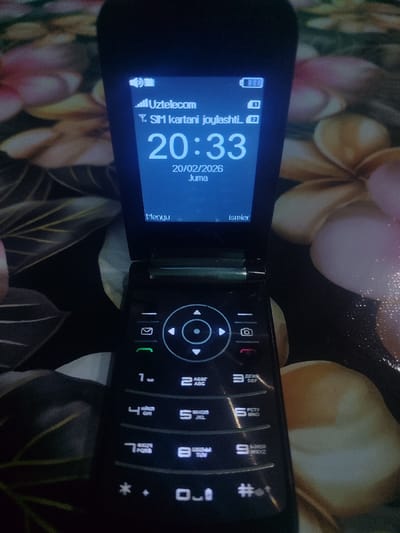The image shows a black flip phone displaying "Uztelecom" and a message saying "Y. SIM kartani joylashti". The time is 20:33, and the date is 20/02/2026, which is a Friday (Juma). Below the date are "Menyu" and "Ismlar" (Menu and Names). The keypad is visible, with labeled keys for digits, letters, and symbols.