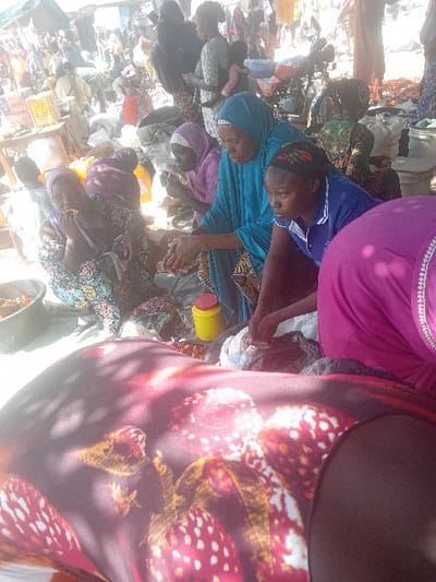 This image depicts a bustling outdoor market scene in Gurku Gwari, Nigeria. The foreground is partially obscured by the back of an individual wearing a dark reddish-brown garment patterned with white and orange-red abstract shapes, upon which shadows are cast.
In the midground, several individuals are engaged in market activities. A woman wearing a teal-blue abaya and matching hijab leans forward, extending both hands over a small display of items on the ground, indicating a transaction or sorting. To her immediate right, a younger woman in a blue polo shirt with a white collar and a patterned headscarf kneels, looking downwards, her hands also interacting with items on the ground. To the teal-clad woman's left, two women are seated on the ground, both consuming food. One wears a purple hijab and patterned dress, holding food to her mouth. The other, further left, is in a light blue patterned dress and headscarf, also eating from her hand.
A bright yellow container with a red lid is visible on the ground near the central group. To the far left, a dark bowl contains reddish-brown material, possibly food or spices. Various market goods, including bags of produce or dry goods, are spread on the ground and on makeshift tables.
The background reveals a crowded market environment with numerous additional individuals standing and moving between stalls. A person carrying an infant on their back is visible in the upper-left background. A motorcycle with a blue seat cover is parked in the upper-right background. The ground appears to be unpaved, likely dirt. Overhead, elements suggest temporary market coverings or canopies, creating dappled sunlight and shadows across the scene. The lighting indicates a bright, sunny day.