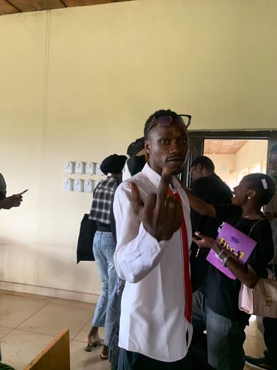 The image depicts an indoor scene, possibly within a building in Farin Yashe, Nigeria. In the foreground, a young Black man wearing a white collared shirt and a red tie is the central figure. He is looking towards the camera and gesturing with his right hand, showing his middle finger. He wears glasses perched on top of his head and has a braided hairstyle. To his right, a young Black woman is visible, holding a purple book titled "Five in 1." She is wearing a black top and a necklace, and her hair is adorned with a pink clip. Behind them, other individuals are present, some of whom are partially obscured. In the background, there are doors leading to another room with what appears to be a tiled ceiling and fluorescent lighting. On the wall to the left, there is a series of light switches or control panels. The overall environment appears to be informal, possibly a community center or educational facility. The lighting is natural and appears to be coming from overhead and possibly from the adjacent room.