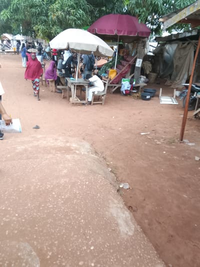 The image depicts a bustling outdoor market scene in Bida, Nigeria. In the foreground, a person in a red headscarf and patterned skirt walks towards the right, carrying a tray of items on their head. To their left, a person in a purple headscarf sits on a wooden stool.
Further into the scene, under a large, white umbrella, several people are gathered around a wooden table. One individual, wearing a light-colored shirt and pants, sits at the table. In the background, there are various market stalls with goods displayed, including what appears to be vegetables and other food items. Two large umbrellas, one white and one burgundy, provide shade over the stalls.
The ground is predominantly red earth, typical of many Nigerian landscapes. In the middle ground and background, several motorcycles are visible, suggesting transportation within the market. The overall environment appears to be a busy, open-air marketplace with a mix of vendors and shoppers interacting.
