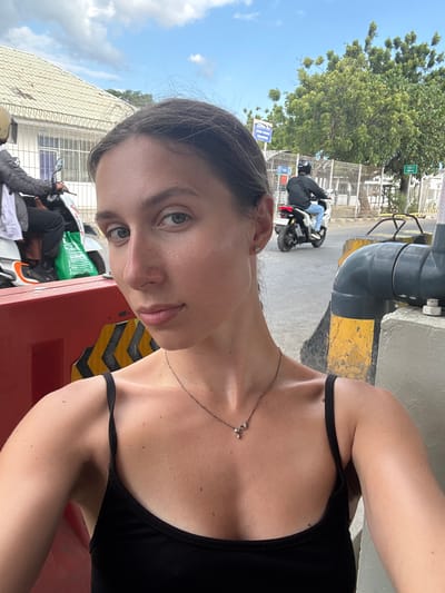 An outdoor, close-up, selfie-style image shows a young woman with light skin, dark hair pulled back, and light-colored eyes looking at the camera. She is wearing a black tank top and a thin necklace with a small pendant. Her left shoulder and décolletage are visible, along with a diamond stud earring in her left ear. Behind her, a street scene unfolds. A red and yellow striped barrier is in the foreground. Further back, a person wearing a helmet is riding a scooter with a green bag on the back. Another person on a scooter with a helmet is visible in the middle ground. Buildings and trees line the street, under a partly cloudy blue sky. A sign that reads "ZONA AJ" is visible on a building in the background. A sign pointing to "TOILET" is also visible. The overall impression is a casual snapshot taken in an urban environment in Upt Tambak Sari, Indonesia.