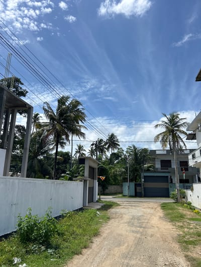 The image depicts a street scene in Kadewatta, Sri Lanka, under a bright blue sky with scattered wispy clouds. A dirt road leads into the distance, flanked by residential buildings and lush tropical vegetation, including several palm trees. On the left side of the road, a partially constructed building with concrete pillars is visible, adjacent to a white painted wall. The right side of the road features multi-story houses with balconies and prominent electrical wiring overhead. Small flags are visible on some of the buildings. The environment appears to be a suburban or semi-rural area.