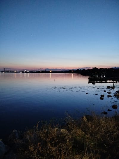 This image captures a twilight scene at the bay of Kazashko, Bulgaria. The sky is a gradient of deep blue at the top fading to lighter blues and pinks near the horizon. A single bright star is visible in the upper left quadrant of the sky. The calm water reflects the sky's colors and the distant lights of an industrial complex on the far shore, characterized by towers and illuminated structures. On the right side of the image, a dark wooden pier extends into the water, with some structures or lights visible on it. The foreground shows a rocky shoreline with dry grasses and vegetation. The overall atmosphere is serene and quiet, indicative of dusk.