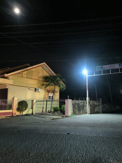 This media file captures a serene night scene in Meuse, Indonesia, illuminated by both natural and artificial light sources.
**Subjects:** The central subjects include a single-story house, a street light, and a road. In the upper left, a bright full moon is visible in the dark night sky, partially obscured by thin clouds. Numerous power lines stretch horizontally across the upper portion of the frame. On the left, a light yellow or tan house with an ornate gable roof and reddish-brown trim stands behind a metal fence and gate. The house features decorative relief work on its gable, an air conditioner unit mounted on a wall, and a distinctive white-framed window. A small palm tree grows next to the house, and a small, round shrub is positioned near the front fence. On the right, a tall street light with a brilliant white-blue glow illuminates the immediate vicinity. Above and behind the streetlight is a structure spanning the road, holding a large sign. A concrete wall and a metal gate are visible to the right of the house. The foreground shows a dark asphalt road with distinct textures, possibly from recent rain or the way the light catches its surface.
**Activity or event taking place:** The scene appears quiet and devoid of any human activity, suggesting a late hour.
**Notable details:** The time of day is clearly night, with the moon providing some ambient light alongside the strong, bright artificial light from the street lamp. The street lamp casts a strong glare, creating a lens flare effect. The architectural details of the house, particularly the intricate relief work on the gable, are notable. The presence of power lines adds to the urban or semi-urban setting.
**Visible text:** The sign above the streetlight clearly reads "Selamat datang di DESA KULU," which translates to "Welcome to Kulu Village," confirming the location context of Meuse, Indonesia. Below this main text, smaller text is visible, including words like "MEUSE..." and "HUT..." (likely an abbreviation for "Hari Ulang Tahun" or anniversary), suggesting information about the area or a local event.