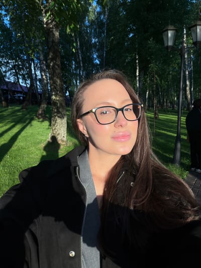 A woman is photographed outdoors in what appears to be a park or wooded area. The background includes trees, grass, and lamp posts. The woman is wearing glasses, a grey top and a black jacket. The photo seems to be taken in Polivanovo, Russia.