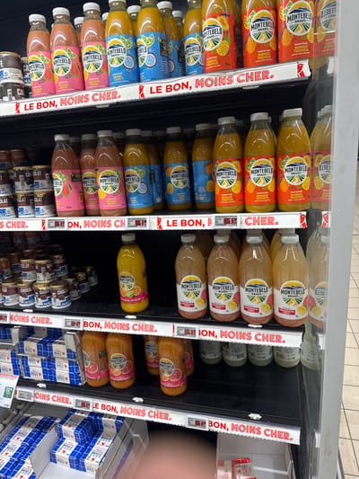 The image shows a refrigerated display case in a supermarket in La Trinité, France, stocked with various beverages and some food items. The shelves are organized with rows of bottles and containers. On the top shelf, there are several bottles of Montebelo smoothies in different flavors, including "Goyave Amour," "Samba Coco," and "Mango Tropico." Below this, on the second shelf, there are more Montebelo smoothies, some in larger 1-liter bottles labeled "Pamplemousse Citron." There are also some smaller containers of what appear to be chocolate products on the third shelf from the top. A price tag with "1.77 €" is visible on the second shelf. Red and white promotional banners with the text "LE BON, MOINS CHER." are placed along the edges of the shelves, indicating a sale. The overall environment is brightly lit, typical of a retail setting. There are no people visible in the image.