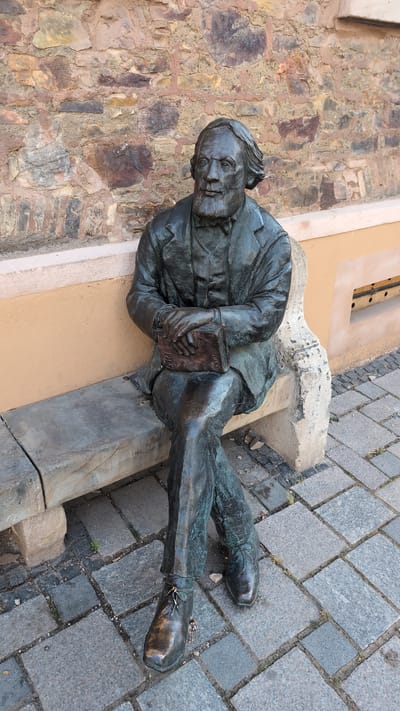 A bronze statue of a seated man is positioned on a stone bench in front of a textured stone wall. The man, depicted with a beard and wearing a suit and bow tie, is holding an open book in his lap. His legs are crossed, and he is wearing dress shoes. The bench is made of rough-hewn stone. The background wall is constructed of irregular stones of varying colors, from brown to tan, with visible mortar. To the right of the bench, a light-colored wall with a small grated opening is visible. The ground is paved with cobblestones. This statue is located on the banks of the Rhine River in Bingen am Rhein, Germany, specifically in front of the "Mäuseturm" (Mouse Tower) museum.