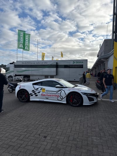The image depicts an outdoor scene at the Sachsenring race track in Hohenstein-Ernstthal, Germany, under a partly cloudy sky. The foreground shows a white Honda/Acura NSX sports car, presented in profile, facing right. The car features a livery with "ADAC Sachsen," "Fahrsicherheitszentrum Sachsenring" alongside an outline of the Sachsenring track, "Sachsenring.de," a black and white checkerboard pattern, and "BRIDGESTONE" branding. It has black multi-spoke wheels with visible red brake calipers.
To the immediate left and slightly behind the sports car, a black scooter is parked. In the left foreground, the back of a person wearing a dark top and light lower garment is partially visible. To the right of the sports car, a male subject in a dark short-sleeved shirt, blue jeans, and white sneakers walks from right to left across the paved surface, which consists of dark gray interlocking pavers.
In the mid-ground, a large white and gray semi-trailer truck is parked, displaying "DEKRA" logos and the slogan "Was die Welt bewegt, machen wir sicher." Further right, a group of approximately five to six individuals is gathered near the truck and a modern, multi-story grey building with a yellow vertical accent panel. One person in this group wears a yellow shirt. A motorcycle is partially visible behind this group on the right. Multiple green "DEKRA" flags and yellow "ADAC" flags are visible on poles throughout the background. The sky above is blue with scattered white cumulus clouds.