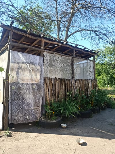 A wooden, open-sided structure resembling a shelter or shack stands outdoors under a clear blue sky. The structure's roof is made of wooden planks and logs. Its walls are partially covered by light-colored, patterned fabric curtains. The front of the structure is bordered by a fence made of vertical wooden sticks. In front of the structure, several old tires have been repurposed as planters, containing green plants. The ground in front of the shelter is dirt and cracked concrete. Two shallow bowls, one with some white residue, are visible on the ground in the foreground. The scene is located in the village of Auly, Dnipropetrovsk Oblast, Ukraine. The overall impression is one of a rustic, utilitarian outdoor space.