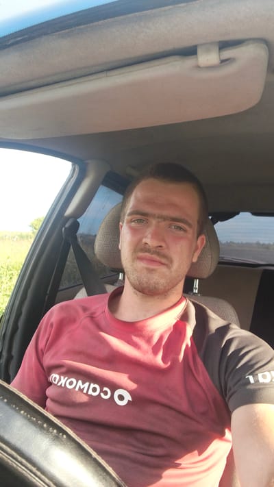 The media file is a selfie taken by a young man sitting in the driver's seat of a car. He is looking directly at the camera with a neutral expression. He has short brown hair and a faint stubble on his face. He is wearing a red and gray t-shirt with white text on the front that appears to be a logo. The interior of the car is visible, including the car's sun visor, the headrest of the passenger seat, and the steering wheel. Sunlight is streaming into the car, suggesting it is daytime and the weather is fair. The background outside the car appears to be a rural landscape with greenery, possibly fields. The scene is set in Kolobovo, Russia.