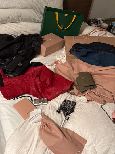 The image depicts a cluttered bed in an indoor environment, likely a domestic setting. The scene includes several high-end fashion items strewn across a white duvet. Noteworthy objects include a Goyard shopping bag, a Dior bag, several Chanel handbags, and a Miu Miu item, as well as several pieces of clothing. The overall impression is one of disorder and luxury. Location is unknown. There is no indication of the location being Chula Vista, United States of America. No people are visible.