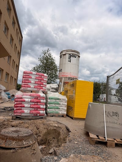 The image shows a construction site in Příbram, Czechia, on a cloudy day. In the foreground, a large concrete manhole cover is visible, partially submerged in dirt. Behind it, stacks of construction materials are arranged. To the left, there are two large stacks of red and white bags, likely containing building supplies, wrapped in plastic. To the right of these, a stack of green and white bags is partially visible. Further back, a large, cylindrical white silo with black lettering is present, and next to it is a tall, bright yellow stack of materials, possibly insulation or blocks. A concrete barrier is placed on a wooden pallet in the right foreground. In the background, a multi-story building with beige siding and white-framed windows stands to the left. A chain-link fence encloses the construction area on the right, with a white van visible behind it. Trees are scattered throughout the scene. The ground is covered with gravel and dirt.
