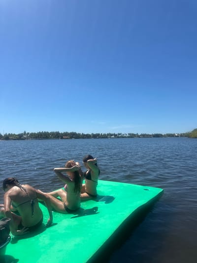 Three women in bikinis are on a green platform on the water. They appear to be shielding their eyes from the sun. The water is dark, with ripples creating texture. A line of trees is visible in the distance across the water. The sky is a clear, bright blue. The platform takes up a significant portion of the right side of the image.