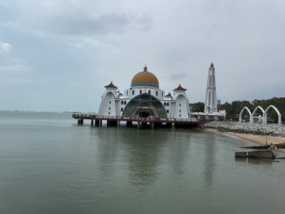 A waterfront mosque, with a large golden dome trimmed in blue, sits on stilts above the water. A white tower and arched walkway connect the structure to the shore. The sky is overcast. A small sandy beach leads up to the walkway, and some people are walking on the beach near concrete blocks. The water is calm and reflects the mosque and sky. A distant landmass is visible on the horizon.