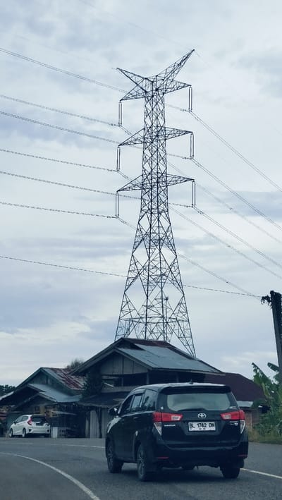 This image captures a black Toyota Innova driving on a paved road, viewed from its rear-right, against a backdrop dominated by a large electricity transmission tower and an overcast sky. The setting appears to be a semi-urban or rural area in Lambarotunong, Indonesia.
The towering power pylon, with its intricate metal lattice structure and multiple overhead power lines, stands prominently in the upper half of the frame. Below the pylon and behind the black car, several weathered buildings with corrugated metal roofs are visible, suggesting local architecture. Another white car, possibly a Honda Jazz/Fit, is parked further down the road on the left side.
The sky is cloudy, indicating an overcast day, perhaps in the morning or late afternoon, with soft, diffused lighting. The road features a white broken line along its curve. The black Toyota Innova has a clear license plate, "BL 1742 DN," with "BL" being a regional code for Aceh, Indonesia. The car's make "TOYOTA" and model "INNOVA" are also visible on its rear. Sparse vegetation, including banana leaves, can be seen on the right side of the frame. The scene portrays a typical day, focusing on infrastructure and local transportation.