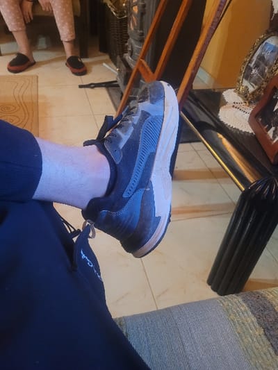 An indoor scene in Κωστήτσι, Greece, depicts a person's lower left leg and foot in the immediate foreground, positioned over a patterned sofa or chair. The individual wears dark blue or black sweatpants with a white logo on the thigh and a dark grey/black athletic shoe with a tan sole. The floor is tiled with light-colored square tiles, partially covered by a light-patterned rug. In the background, two pairs of bare feet wearing red and black slip-on slippers are visible on the floor, suggesting the presence of other individuals. To the right, a dark, ornate structure, possibly a stove or fireplace, stands beside a wooden frame leaning against a warm yellow wall. A dark, reflective table, adorned with a white crocheted doily and two framed photographs (one gold, one reddish-brown), is positioned against the wall. A dark, ribbed decorative object is visible below the table.