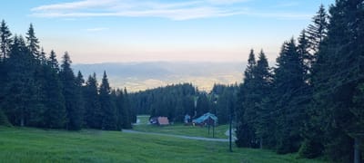 This image captures a serene mountain landscape in Martin, Slovakia, likely during the late afternoon or early evening, given the soft, diffused light and the pastel hues in the sky. The scene is dominated by a lush, green meadow in the foreground, sloping upwards towards a dense forest of tall, dark green coniferous trees.
Nestled within the treeline, a cluster of buildings forms a small settlement. These appear to be rustic mountain lodges or houses, with one prominently featuring a red-tiled roof. A ski lift structure, characterized by poles and wires, is visible on the right side of the image, suggesting the location is a ski resort or a popular mountain recreation area.
Beyond the immediate forest, the landscape opens up to reveal a vast valley. Rolling hills and distant mountain ranges stretch as far as the eye can see, fading into a hazy, atmospheric perspective. The valley floor is dotted with patches of lighter green, possibly fields or smaller clearings, and hints of a town or village can be discerned in the distance.
The sky above is a clear blue, gradually transitioning to softer shades of pink and orange near the horizon, indicative of the approaching sunset. A few wispy clouds add a delicate texture to the upper atmosphere. The overall mood is one of tranquility and natural beauty, evoking a sense of peace and remoteness. There is no visible text in the image.