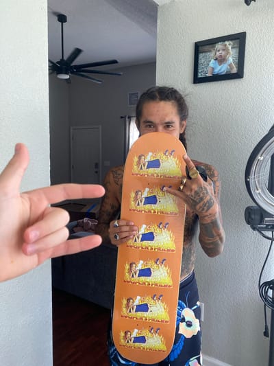 In Elizabeth City, a person with tattoos and braided hair stands in an indoor setting, holding an orange skateboard with a repeating graphic of a woman surrounded by flames and the words "LUCIFER'S FRIEND." The skateboarder is wearing patterned shorts and is adorned with rings on their fingers. A framed picture of a young girl hangs on the textured wall behind them. In the background, a ceiling fan is visible, along with a door and part of a couch. To the left, a hand with two fingers extended forms a 'rock on' gesture. To the right, a ring light setup is partially visible.
