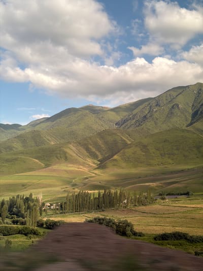 The image displays a wide, sweeping landscape with a dramatic mountain on the left, partially obscured by a vast expanse of white, fluffy clouds against a blue sky. The mountain is covered in lush green vegetation, with visible textures and contours suggesting steep slopes and valleys.
To the right of the mountain, the scene transitions into rolling hills that have been cultivated into agricultural fields. These fields exhibit varying shades of green and brown, with distinct rows indicating farming practices. A line of tall, slender trees runs along a portion of the cultivated area, creating a defined edge between the fields and a small settlement.
The settlement appears to be a cluster of buildings nestled within the landscape, surrounded by more trees and vegetation. This hints at a rural or semi-rural setting. Further to the right, the foreground is dominated by a blurred, dark brown area, which could be a dirt road, a fallow field, or the edge of another cultivated area.
The lighting suggests it is daytime, with sunlight casting shadows and highlighting the textures of the land and clouds. The weather appears fair with scattered clouds. The overall impression is one of a vast, natural landscape with human intervention in the form of agriculture and a small community. The location is identified as Karagai Bulak, Kyrgyzstan, suggesting a mountainous and possibly agricultural region in Central Asia. No visible text is present in the image.