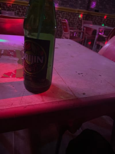 A partially consumed green glass bottle of "ORIGIN" brand spirit mixed drink, containing a light green straw, rests on a light-colored, scuffed table. The table surface exhibits minor marks and is predominantly illuminated by red and purple ambient lighting, casting reflections. The image is set within an indoor establishment in Mape, Nigeria, characterized by a patterned wall with a gold trim visible in the background. Multiple red-toned chairs and additional tables are discernible in the blurred mid-ground, suggesting a bar, club, or restaurant environment. Distant, colored lights (red, green, purple) contribute to the overall dim and vibrant atmosphere. No individuals are clearly visible, though a dark form partially occupies the lower right edge of the frame, indicating the immediate presence of a seated person.