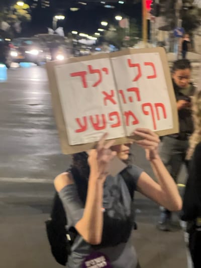 The image depicts a nighttime urban scene in West Jerusalem, Israel, featuring an individual holding a protest sign. The central figure, with dark curly hair, wears a grey short-sleeved top and a dark backpack, with a purple tag attached to their left side. Their face is obscured by a large, open-book-shaped sign constructed from cardboard with white pages. The sign displays Hebrew text in red: "כל ילד הוא חף מפשע," which translates to "Every child is innocent of crime." The individual holds the sign with both hands. To the right, another person with dark hair is partially visible, appearing to engage with a mobile device. The background reveals a wet asphalt road reflecting the bright headlights of multiple vehicles, indicating active traffic. Streetlights are visible further down the street, and a red traffic signal is present on the upper right of the frame. The environment is consistent with a city street after dark.