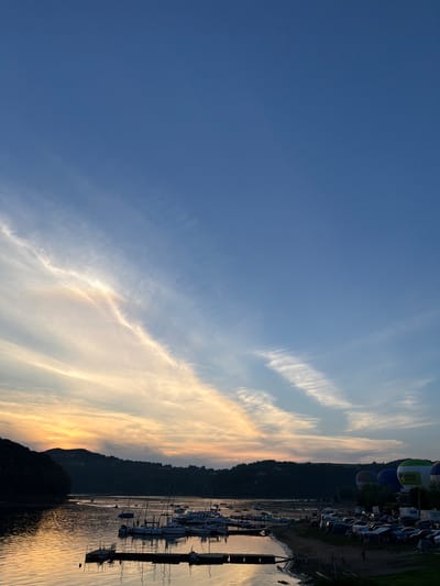 The image depicts a sunset scene at Gródek nad Dunajcem, Poland, on the Rożnowskie Lake. The sky is a gradient of deep blue at the top, transitioning to lighter blues and soft oranges and yellows near the horizon, with wispy clouds streaking across.

The foreground features a body of water, likely the lake, with its surface reflecting the colors of the sky. Several moored sailboats are visible, their masts creating a delicate silhouette. A wooden pier or dock extends into the water. Along the shoreline, a collection of tents and parked vehicles, including cars and a camper van, suggest a temporary encampment or event. Several hot air balloons are also present, some inflated and standing upright, adding a colorful element to the scene.

In the background, rolling hills covered in dense dark foliage form the outline of the landscape. The overall impression is one of a serene evening by the water, with signs of outdoor recreation and possibly a festival or gathering. The lighting is characteristic of dusk, with soft, diffused light casting long shadows. No specific actions or interactions between people are clearly discernible.