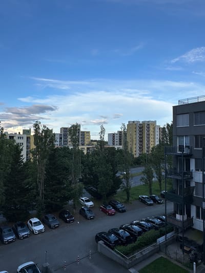 This is an eye-level shot taken in Rüsselsheim am Main, Germany. The image captures a scene featuring a row of high-rise apartment buildings of varying heights, distinguished by their light yellow and off-white facades. These buildings are partially obscured by dense, mature trees with full canopies. A street runs adjacent to the buildings, lined with parked cars of various colors. The sky is a clear, bright blue, with wispy white clouds. The architectural style of the buildings suggests a modern or post-modern design. A portion of a building with balconies is visible in the upper section of the frame.