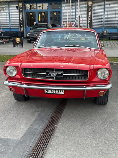 A red vintage Ford Mustang is parked on the side of a road in Wetzikon (ZH), Switzerland. The car faces the viewer, showcasing its front grille, headlights, and a license plate that reads "ZH-761335". The reflection on the car's hood suggests an urban environment. To the left of the vehicle, there's a blue building marked with "Vettiger Kunden" signs. A black Mercedes is parked behind the Mustang. The foreground includes a section of sidewalk and a metal street drain. The image captures an outdoor scene during daylight.