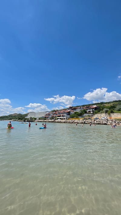 This is a vertical, eye-level shot of the sea at Topola, Bulgaria. The water is clear and calm, with sunlight creating ripples on the surface and illuminating the sandy bottom. Several people are visible in the water, some on paddleboards and others swimming. In the background, a rocky shoreline separates the sea from a hillside dotted with buildings and trees. The sky above is a vibrant blue, with scattered white clouds. The buildings appear to be modern hotels or apartment complexes, constructed with white facades and red-tiled roofs. The scene conveys a sense of a peaceful summer day at the beach.