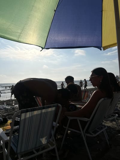 The image depicts a beach scene in Montañita, Ecuador (latitude: -1.825, longitude: -80.754). A group of four people are gathered under a colorful beach umbrella, sitting in chairs. One person, a man, is bending over, seemingly looking at something on the ground. Two people are sitting in chairs, and one woman is sitting in a chair, looking to the right of the image. The beach is sandy, and the ocean can be seen in the background. There are a few people in the distance, some standing on the beach and some wading in the water. The image was taken during the day, and the sun is visible through the umbrella. The scene is casual and relaxed. The overall ambiance is sunny and warm.