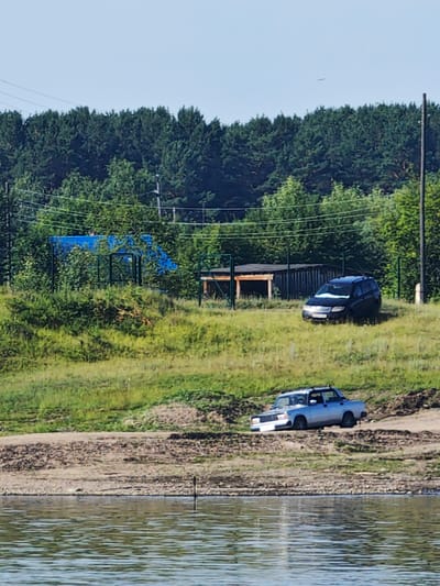The image depicts a rural outdoor scene with a body of water in the foreground, likely a river or lake, with visible ripples on its surface. Beyond the water, a dirt bank leads up to a grassy slope. Further up the slope, a light blue sedan is parked at an angle, partially on the dirt and partially on the grass. Behind it, a dark-colored van is parked on the grassy area. A fence line runs across the middle ground, with a blue tarp covering a structure behind it. A wooden shed or shelter is visible within the fenced area. The background consists of a dense line of green trees, with a clear sky above. The image appears to be taken during daylight.