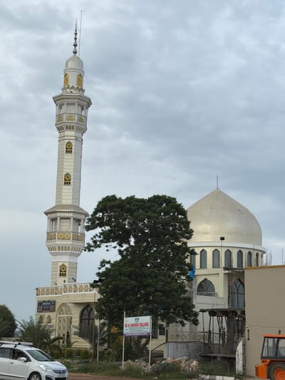 This is a full shot, exterior, vertical, eye-level photograph of a mosque and a junior college in Balanagar, India. The mosque features a tall, white minaret with gold accents and a dome. The junior college is adjacent to the mosque, and construction is visible on a building to the right. A white car is parked in the foreground, and a loader is visible on the far right. The sky is cloudy.