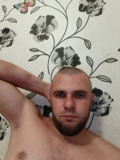 A man with a shaved head and a beard is lying on his back, with his right arm raised behind his head. He is wearing no shirt. The background is wallpaper with a floral pattern. The scene appears to be indoors. The location is the city of Pishchane, Ukraine.