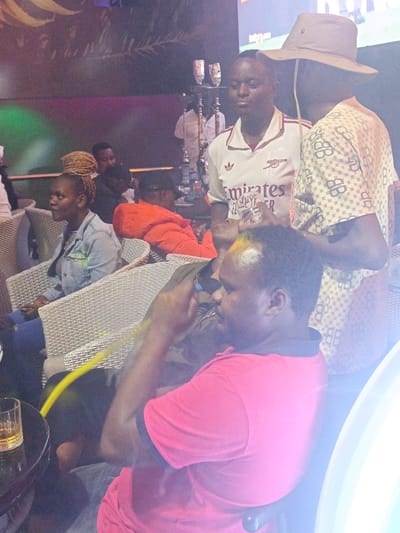 The image depicts an indoor scene in an establishment within the city of Syokimau-Mulolongo ward, Kenya. A group of people are present, engaged in various activities. A man in a pink shirt is seated, holding a yellow hose, likely part of a hookah setup. Another man wearing an Arsenal football jersey stands nearby, while a third man in a patterned shirt and hat is partially visible. A woman with braided hair is seated in a wicker chair, and other patrons are visible in the background. There's a glass with a liquid inside on a table next to the seated man. A hookah is visible in the background. The environment appears to be a bar or lounge, with dim lighting and modern decor.