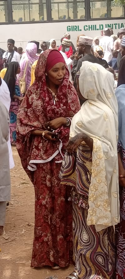 The scene depicts an outdoor gathering in Sani Mai Nagge, Nigeria, with a crowd of individuals, predominantly adults, standing on unpaved dirt ground. In the immediate foreground, two women are engaged in conversation. The woman on the left wears a maroon headscarf and a matching maroon dress adorned with a red and beige floral pattern. She has a gold necklace with a prominent pendant, gold bracelets, a watch on her left wrist, and dark henna or nail polish on her fingernails. Her mouth is open as if speaking, and she holds a dark object, possibly a phone, in her hands. The woman on the right is partially obscured but is visible wearing a cream-colored head covering with gold embroidered floral designs and a patterned skirt featuring swirling lines of purple, brown, and yellow. Her right hand and forearm display extensive dark henna designs.
In the mid-ground and background, numerous other individuals are visible, many wearing traditional attire such as head coverings for women and kufi hats for men. Behind the crowd, a large white bus is partially visible, with multiple windows and the green text "GIRL CHILD INITIATIVE" clearly printed on its side. The overall environment suggests an event or public assembly under daylight conditions.