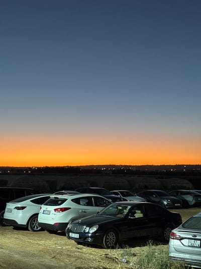 The image captures a parking lot scene at dusk in Um al Amad, Jordan. A row of cars, predominantly white and black, are parked on a dirt and gravel surface. The cars include a white Hyundai, a black sedan, and a silver SUV with license plate "47 49714". The sky displays a gradient of colors, transitioning from a deep blue at the top to a vibrant orange and yellow near the horizon, indicating sunset. In the background, the outlines of buildings and what appear to be greenhouses are visible, with scattered lights suggesting an inhabited area. The overall mood is serene, illuminated by the fading natural light and the artificial lights of the distant settlement. The time of day is clearly dusk, with the weather appearing clear.