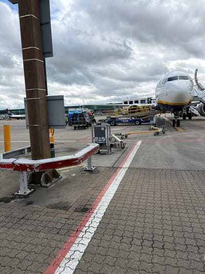 This image captures an active airport tarmac scene in Uttlesford, United Kingdom, under an overcast sky. The primary subject is a Ryanair Boeing 737-800 aircraft, identifiable by its distinctive blue and yellow livery and "RYANAIR" text visible on the fuselage. The aircraft's nose section, including the cockpit windows and the "BOEING" logo, is prominent, with the registration "EI-DSB" faintly visible on its nose.
The aircraft is actively being serviced, surrounded by various pieces of ground support equipment. A blue mobile passenger stairs unit, branded with "BLUE HANDLING" and "SAS," is positioned near an aircraft door. A yellow and black baggage loader or conveyor belt system is extended towards the aircraft's cargo hold, suggesting ongoing loading or unloading operations. Another blue vehicle, also bearing "SAS" branding, is seen near the aircraft. Several ground crew personnel are visible around the equipment, performing their tasks.
In the foreground, a weathered red and white painted metal barrier is positioned on the right, while a large, possibly rusty, metal beam structure stands on the left. The ground is paved with interlocking grey blocks, featuring distinct white and red painted lines that mark boundaries on the tarmac.
The weather is cloudy, with a dense, grey sky suggesting an overcast day. The lighting indicates it is daytime. The scene depicts typical ground operations at an airport, showcasing the coordination of aircraft, ground equipment, and personnel.