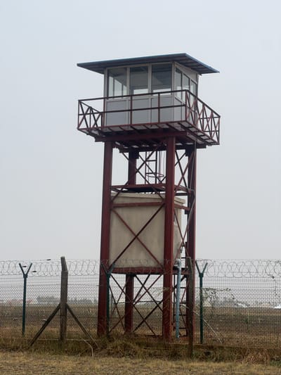 The image depicts a guard tower located next to a wire fence topped with barbed wire. The tower is constructed of red metal beams and has a glass-enclosed cabin at the top. The cabin has a blue roof and a small observation deck. The ground around the tower is dry grass and weeds. The sky is overcast. No people are visible. The location is stated to be Haifam, Nigeria.