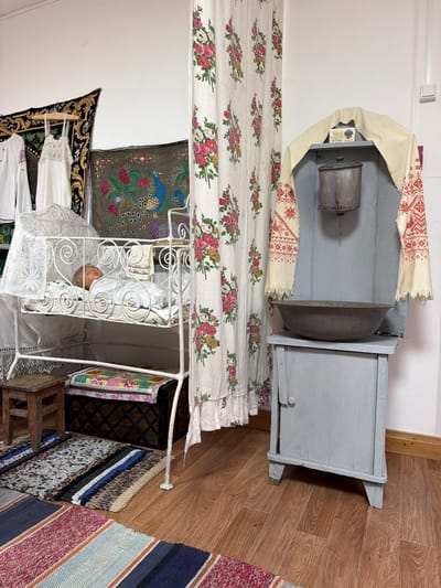 This image depicts an interior scene, possibly a museum exhibit or a preserved historical room, in Ust'-Kabyrza, Russia. The focal point is a white, ornate metal crib containing a doll dressed in white pajamas. The crib has a lace canopy. To the left of the crib, a small wooden stool sits on a woven rug. Behind the crib, a dark wooden chest is visible, topped with a colorful quilted cushion.

To the right of the crib, a tall, pale blue wooden cabinet stands against the wall. It has a small door at the bottom and a large metal basin on a shelf at the top. Above the basin, a metal pot hangs, and a small box labeled in Cyrillic, likely soap, is placed on a piece of fabric. Draped over the top of the cabinet is a cream-colored textile with red embroidered patterns, possibly a shawl or towel.

A floral-patterned curtain, featuring pink and red roses, hangs between the crib and the cabinet, creating a division in the space. On the wall behind the crib and cabinet, several decorative textiles are displayed. One is a dark tapestry with gold and black patterns, and another is a green fabric with embroidered peacocks and floral motifs. Two white garments, one with red embroidery on the sleeve and the other a white lace dress, hang on the left side of the scene.

The floor is made of wooden planks, and several woven rugs are laid out. The overall impression is of a domestic or storage setting, with elements suggesting traditional Russian or Slavic culture. The lighting appears to be artificial, illuminating the objects evenly.