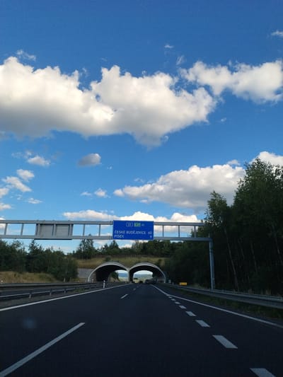 The image depicts a highway scene in Mladotice, Czechia. In the foreground, a black asphalt road with white lane markings stretches into the distance. On the right side of the road, a metal guardrail runs along the edge. Above the road, a blue highway sign indicates "ČESKÉ BUDĚJOVICE 60" and "PÍSEK 9". In the background, two arched tunnels emerge from a hillside covered in trees and grass. The sky is a vibrant blue with scattered white clouds. The overall environment is rural and appears to be during daylight hours. There are no people or specific actions captured in the image.