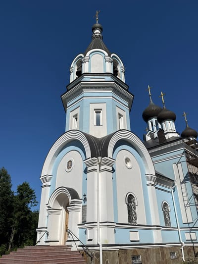 This is a low-angle, full-shot photograph of the Church of the Smolensk Icon of the Mother of God in St. Ozerki, Russia, under a clear blue sky. The church is painted in alternating bands of white and pale blue with white decorative trim. A prominent bell tower with a dark, conical roof topped by a golden cross rises on the left side of the image. Below the bell tower, a series of arched white structures with dark roofs are visible, framing entrances and windows. To the right of the bell tower, three dark, onion-shaped domes, each topped with a golden cross, are part of the main church structure. The building's facade features arched windows with intricate metal grates. At the base of the church, a stone foundation is visible, and a set of red-tiled stairs leads up to an arched entrance. Trees are present on the left side of the frame, bordering the church property. Construction scaffolding is partially visible on the right side of the building. No people are present in the image.